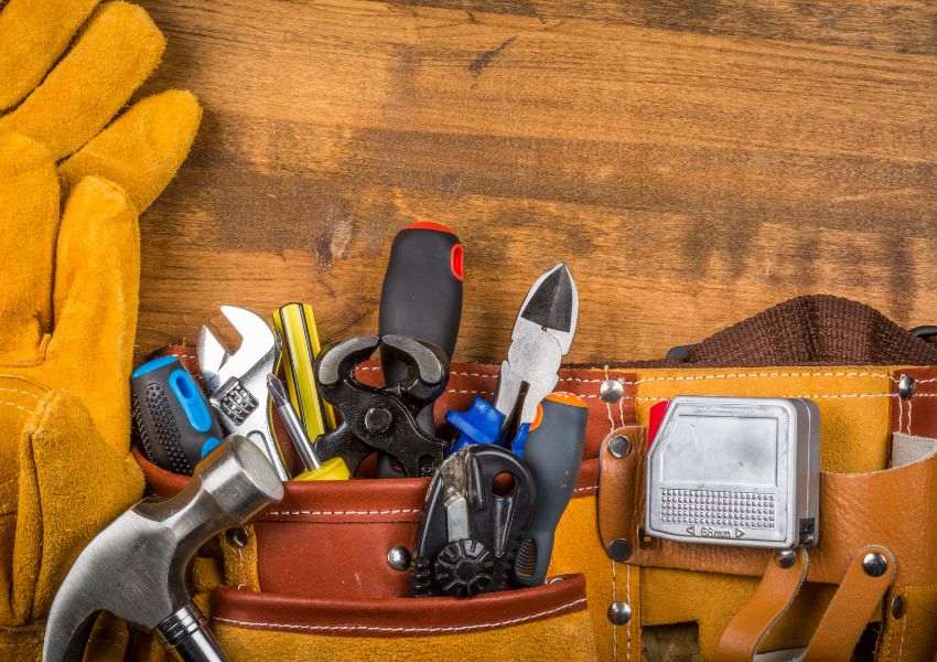 A light brown leather tool belt lies on the hardwood floor, filled with a wrench, hammer, and screwdriver, among other contractor tools.